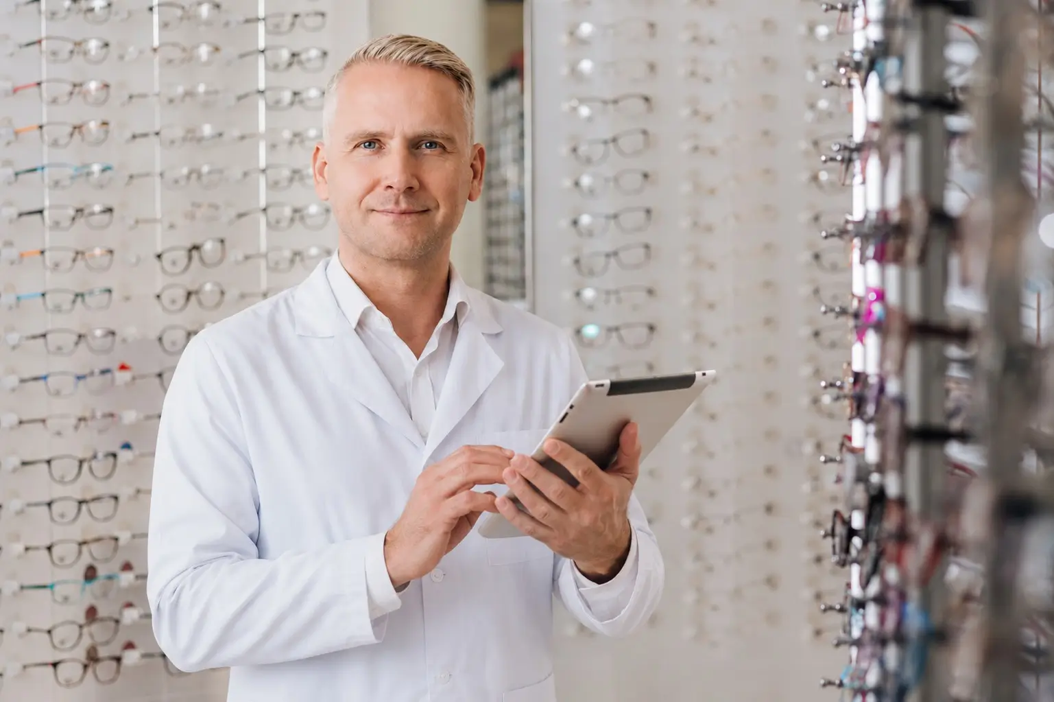 Optometrist in a white coat holding a tablet, standing among shelves of eyeglasses in a brightly lit eyewear shop